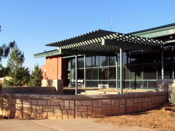 Many Farms High School. School building with green pergola and small stone wall surrounded by desert sand and pine trees. 