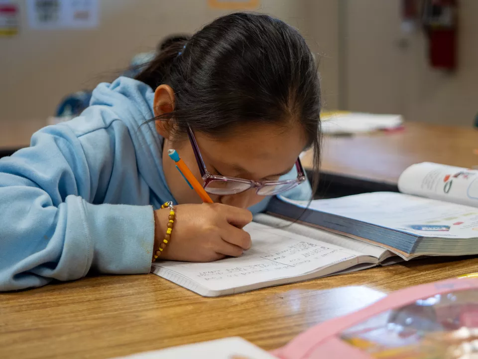 A female student sits with her nose in a book an writes with a pencil on the piece of paper in front of her.