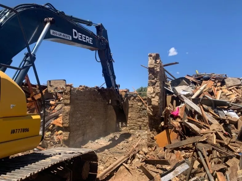 Demolition under way at Western region BIE-funded school. Yellow construction vehicle demolishes deteriorated, brick structure. 