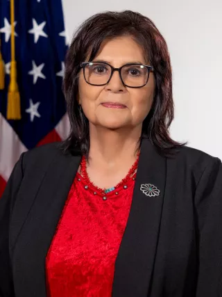 A headshot of a woman with short brown hair in a black blazer and red top in front of an American flag.