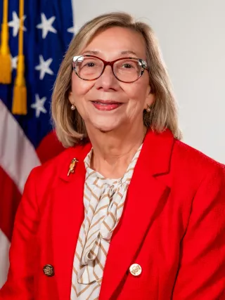 A headshot of a woman wearing glasses, a red jacket, and a beige top in front of an American flag.