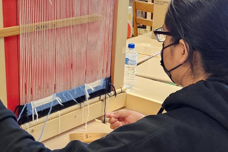 A student sits in front of a traditional loom and learns how to weave.