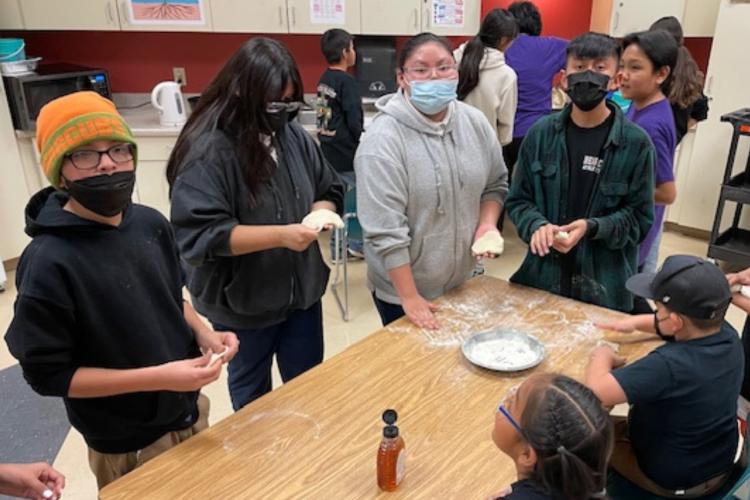 Students stand around a table working with flour and learning to make fry bread.