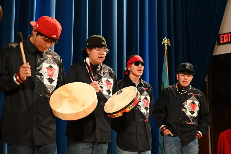 Four male teenagers stand in a row in front of a blue curtain and hold and play their hand drums.