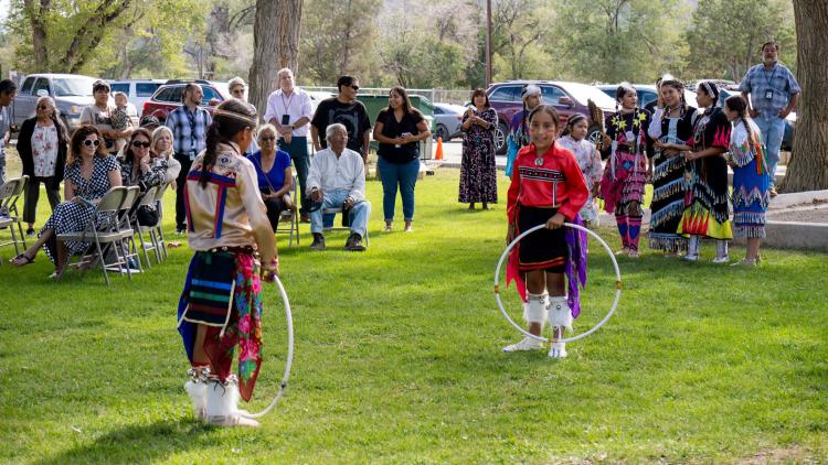 Two students in regalia prepare to hoop dance on a bed of green grass in front of others.