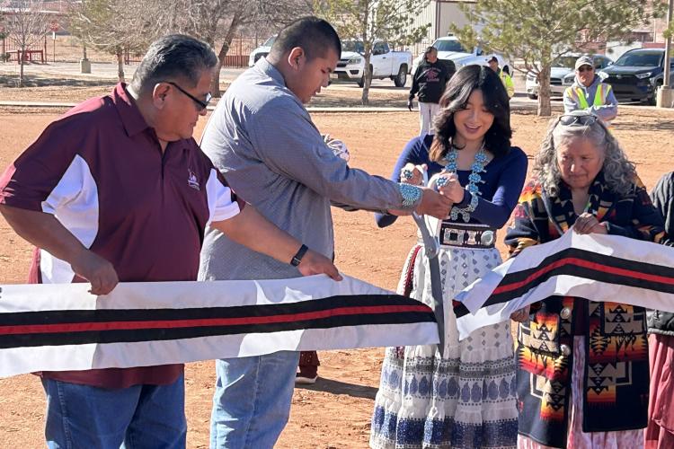 A line of people cut a wide ribbon with a giant pair of ceremonial scissors at a groundbreaking ceremony.