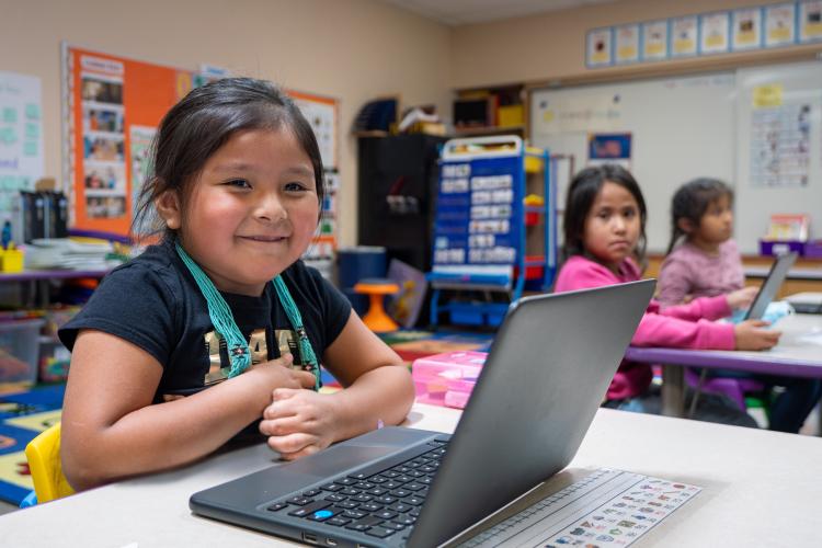 An elementary aged student sits at her laptop and smiles while sitting at her desk.