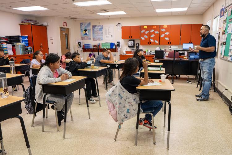 A teacher stands at the front of a classroom facing his students as they sit at their desks.
