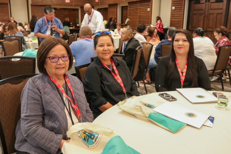 Three women with brown hair in business casual attire sit at a circular table in a conference room.