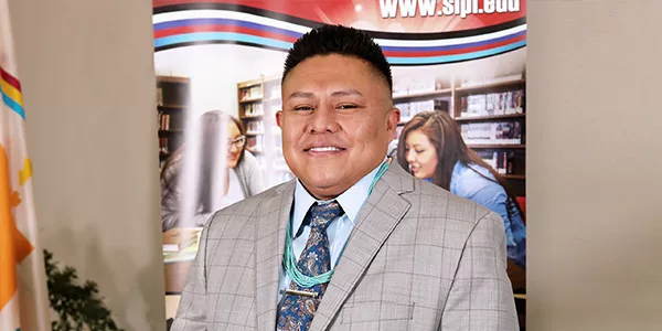 Derrick Shirley, Native American male, smiles for photo in a gray suit, and blue tie. 