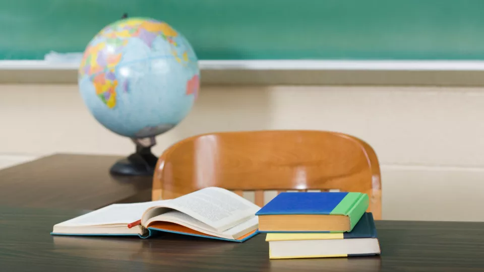 A wooden chair is placed a ta desk that has three books on it with a globe in the background.