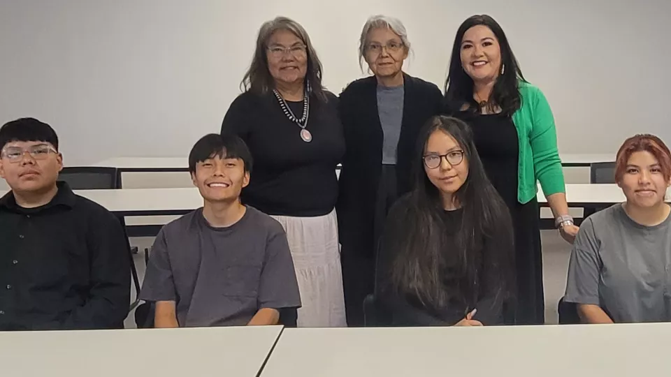 A group of students and their teachers pose for a photo will sitting and standing behind a table.