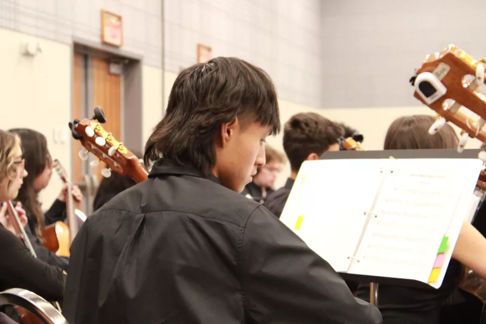 A student in a black shirt plays a guitar while reading sheet music during a performance, surrounded by other musicians in a formal setting.