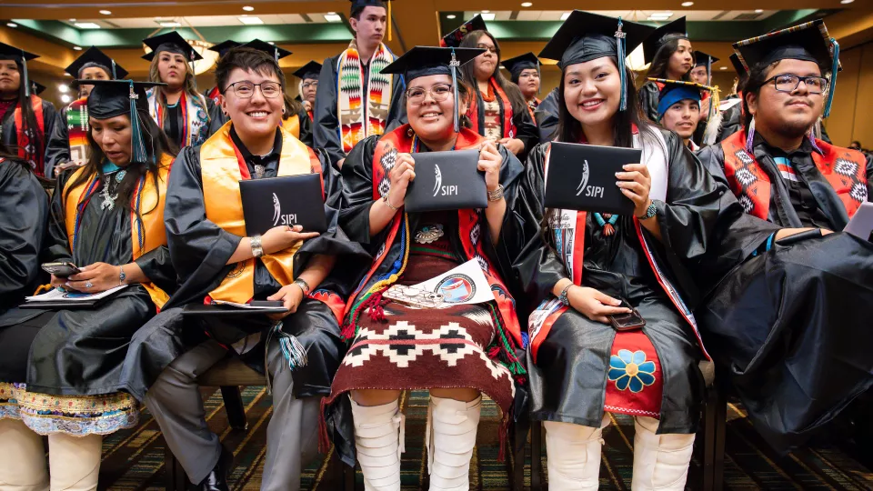 A group of students sit at a graduation ceremony in their caps and gowns, holding up either diploma casings.