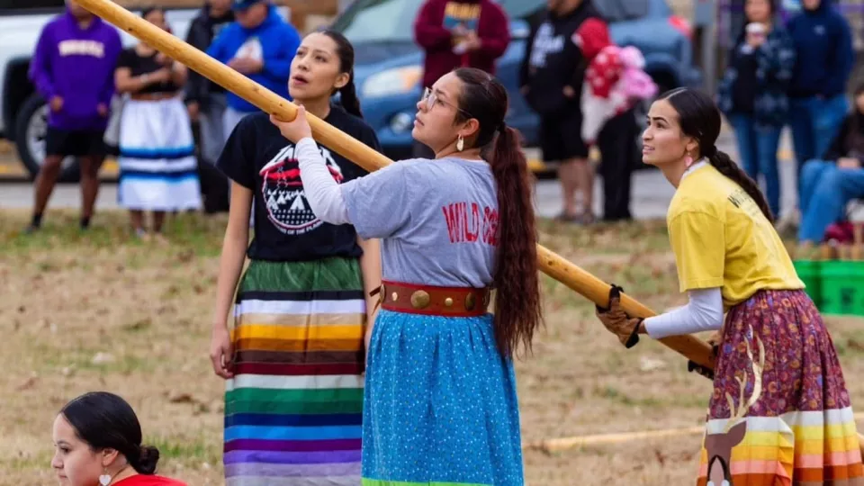 Three women in ribbon skirts hold up a long wooden pole while putting up a teepee.