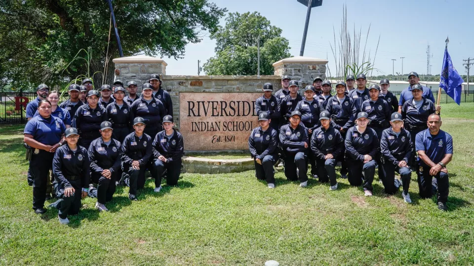 A group of 28 students in cadet uniforms stand and kneel in three lines for a group photo in front of a sign that reads "Riverside Indian School."