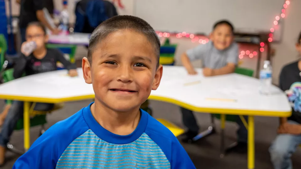 A boy stands smiling in a classroom in front of other students.