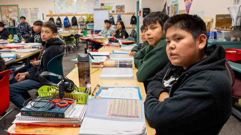 Students sit at their desks, looking toward their teacher at the front of the classroom.