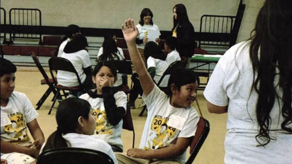 A student raises their hand while sitting with others at a round table.