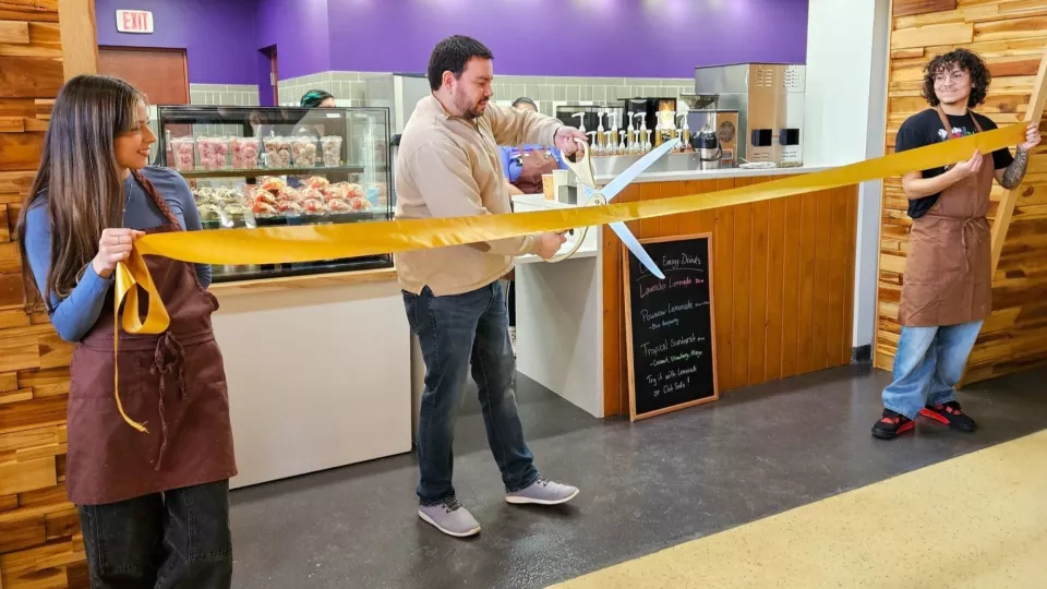 Two people in barista aprons hold up a gold ribbon while a man with giant silver scissors cuts the ribbon in half.