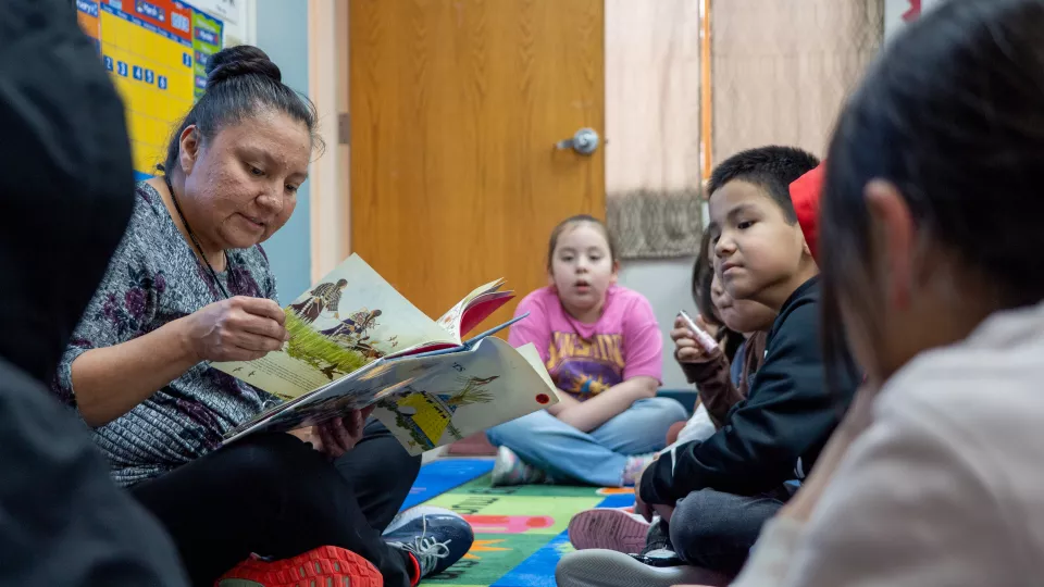 A teacher sits on the ground with her students in a circle and reads to them.