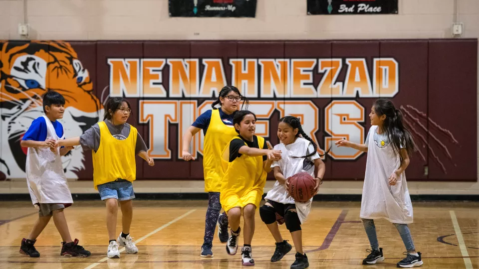 Six students, three in yellow jerseys and three in white, play basketball inside a gym.