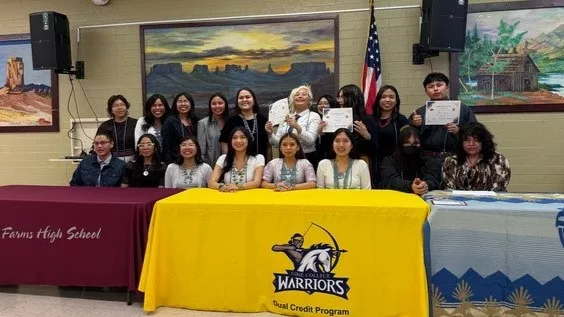 Two rows of students sit and stand behind a table and pose for a photo with a few of them holding up certificates.