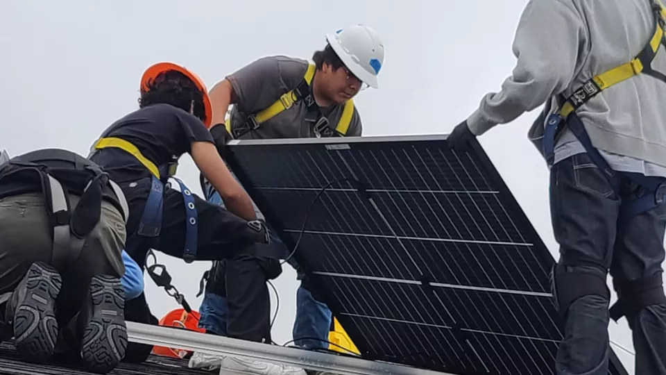 Three people in construction equipment install a solar panel on a roof.