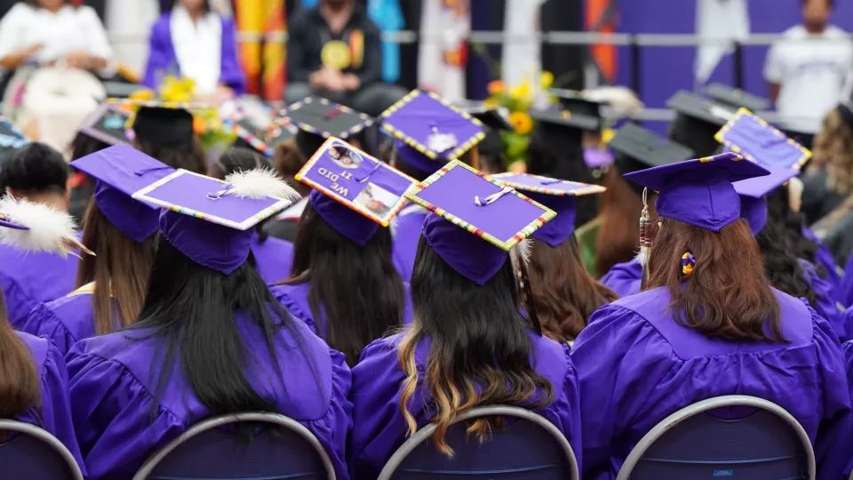 A group of students in purple graduation caps and gowns sit forward listening at a commencement ceremony.