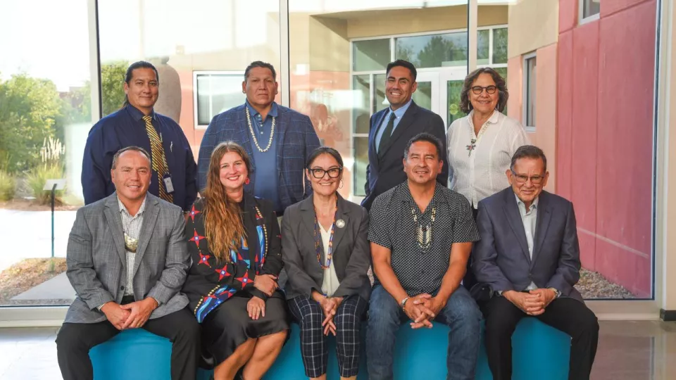 A group of nine adults sit in two lines and pose for a photo outside a building on a school campus.