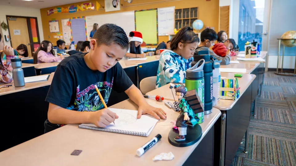 A classroom filled with Native American students engaged in drawing or writing at their desks. In the foreground, a boy wearing a black T-shirt concentrates on his sketch in a spiral notebook. His desk features personal items, including water bottles and a figurine of a character from the anime Demon Slayer. Other students are focused on their work, and colorful decorations and educational materials cover the classroom walls. The atmosphere is creative and focused, with natural light streaming in through la