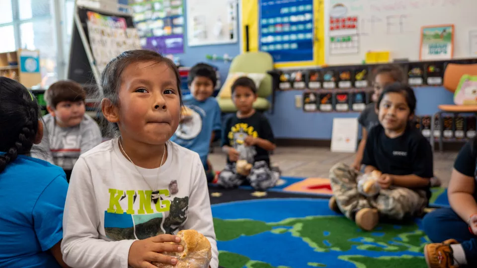 A young Native American girl with dark hair tied back sits on a colorful classroom rug, holding a sandwich and looking off to the side with a slight smile. She wears a white long-sleeve shirt with "KING" and a panther graphic. Other children, also eating, sit around her in a semi-circle, some slightly out of focus. The classroom has bright decorations, including a whiteboard with writing, colorful posters, and an alphabet chart. The setting appears warm and engaging, suggesting a school or early learning en
