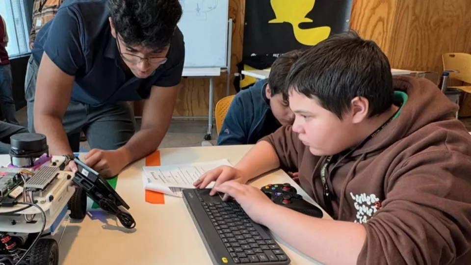 Two students work with an adult at a computer to program a race car robot sitting in front of them.