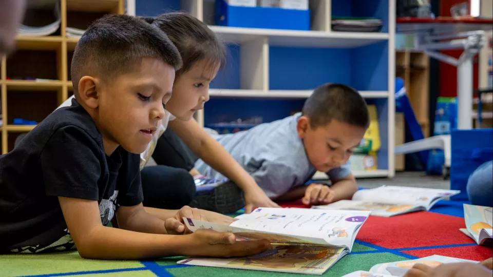 A group of elementary school students lay on their stomachs on a rug in a classroom and read.