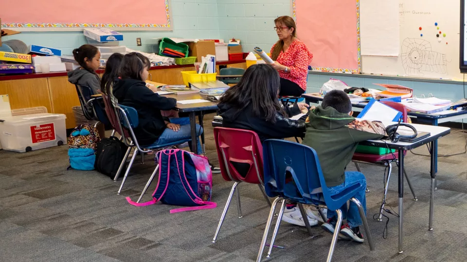 A group of six students sit at two tables facing their teacher who is reading to them.