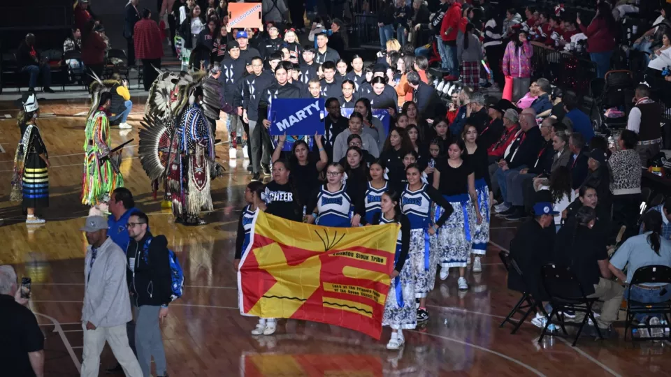 A large group of high schoolers walk in a large parade line onto a basketball court behind the Yankon Sioux Tribe flag led by the school's cheerleaders.
