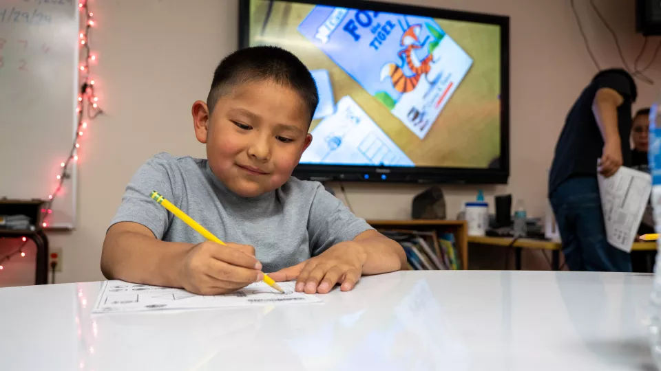 An elementary school student sits at a desk and fills out a worksheet with a pencil.