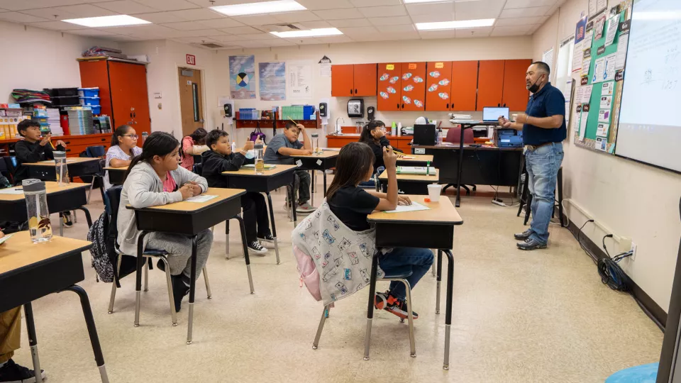 A classroom of students sit at their desks and face forward to listen to their teacher at the front of the classroom.