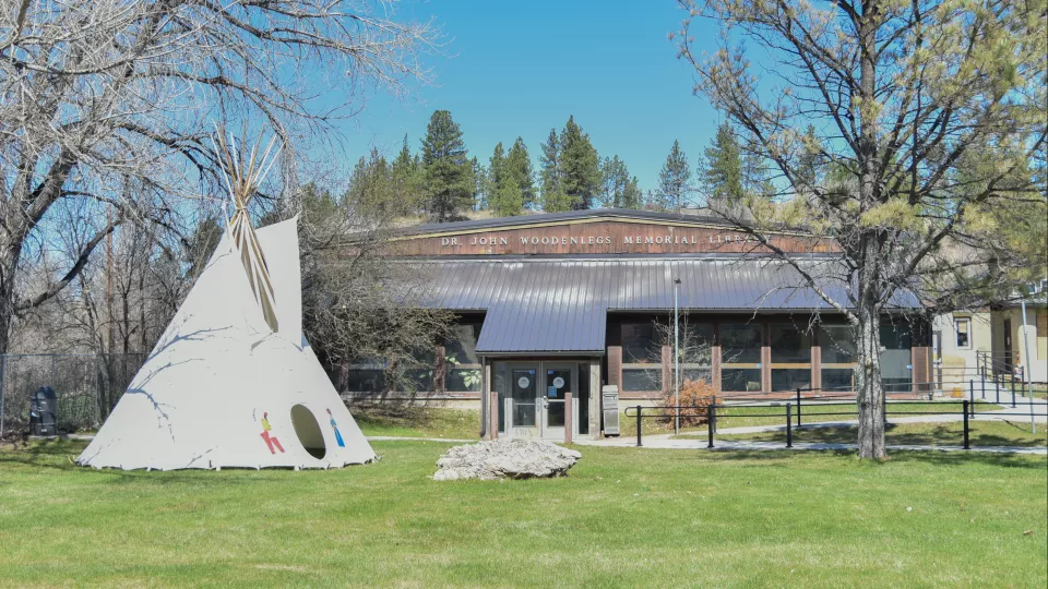 A teepee sits on a field of grass in front of a library surrounded by trees.