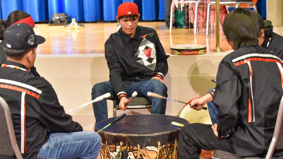A group of male teenagers sit around a powwow drum playing and singing a song.