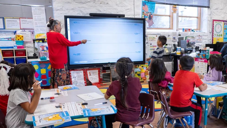 A teacher in a red sweater stands at a smartboard while a class of students faces it and listens.