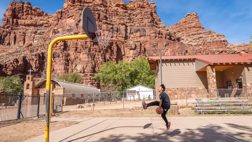 A student plays basketball on an outside court surrounded by rocky mountains.
