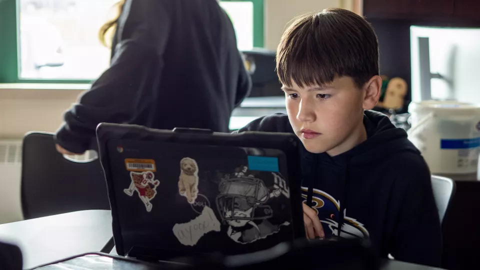 A student in a black hoodie sits in front of a laptop at a desk looking at the screen.