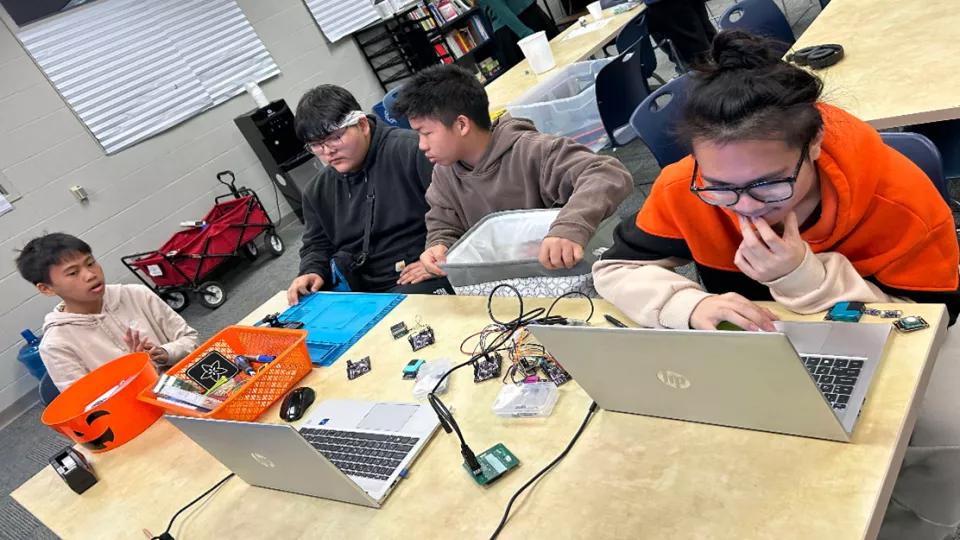 A group of four students sit at a table with laptops and electronic equipment.