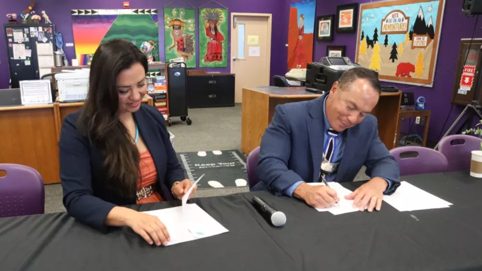 Two people sit at a table in a classroom and sign documents.