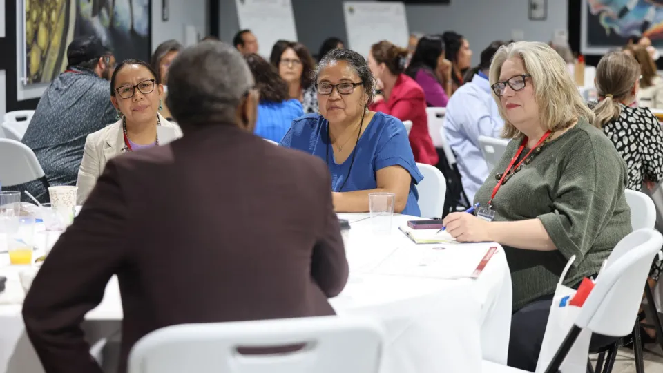 A group of adults sit at a round table with a white table cloth and three women look at a man speaking.