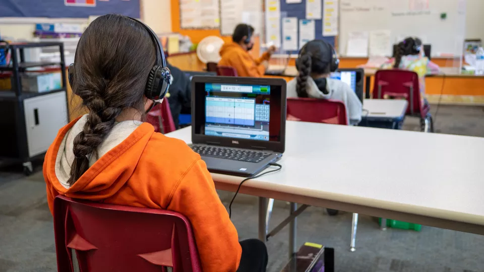 A female student with a long brown braid sits at a laptop and wears headphones.