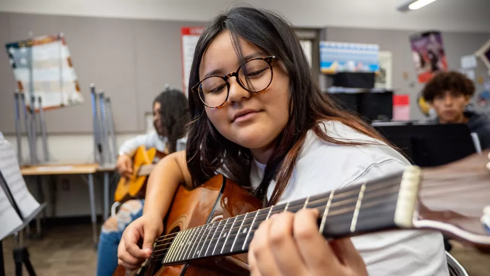 A high schooler with long, brown hair and glasses plays the guitar.
