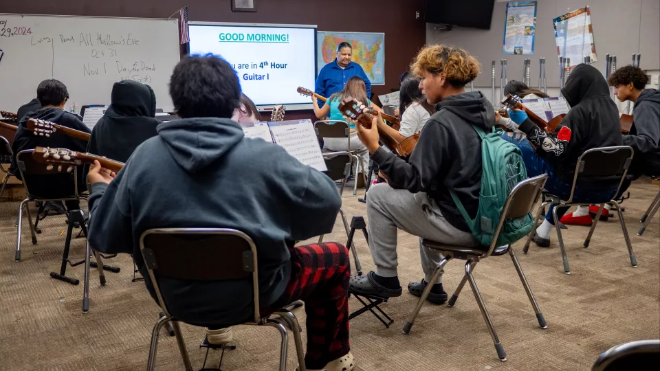 Students sit and learn guitar in a classroom.