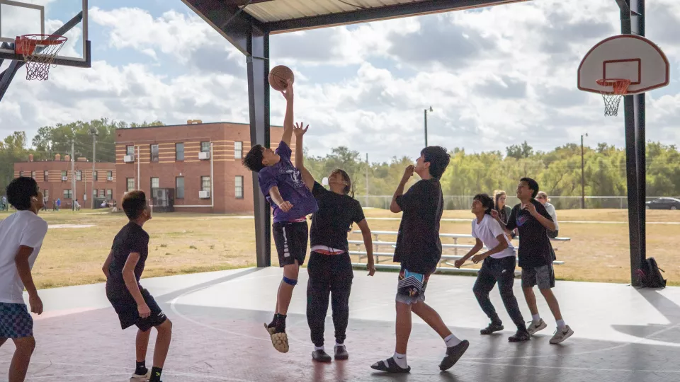 A group of male students plays basketball underneath a covered court at lunchtime.